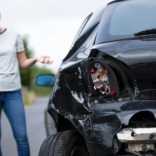 A woman on the phone stands beside a car with visible rear-end damage after an accident.