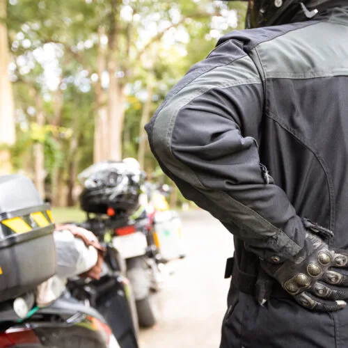Motorcyclist holding their back in pain while standing near a parked motorcycle on the side of the road.
