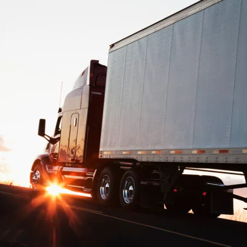 Silhouette of a commercial truck driving on a highway at sunset, with sunlight shining through the truck's wheels.